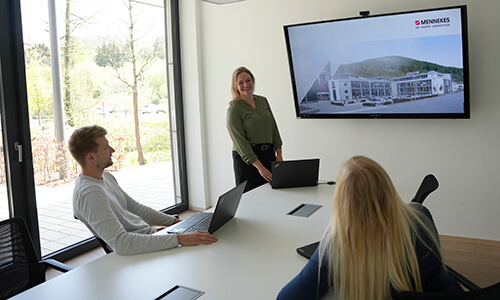 Drei Personen in einem Konferenzraum, zwei sitzen an einem Tisch mit Laptops und eine steht vor einem großen Bildschirm, der eine Präsentation zeigt.