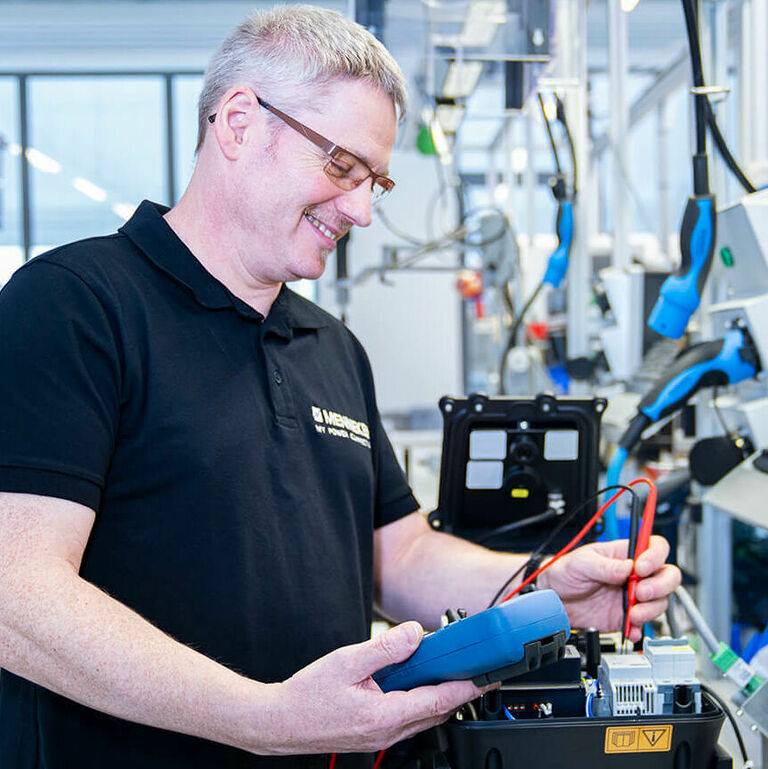 Ein Techniker in einem modernen Labor prüft eine elektronische Schaltung mit einem Messgerät. Er trägt ein schwarzes Polohemd und eine Brille. 