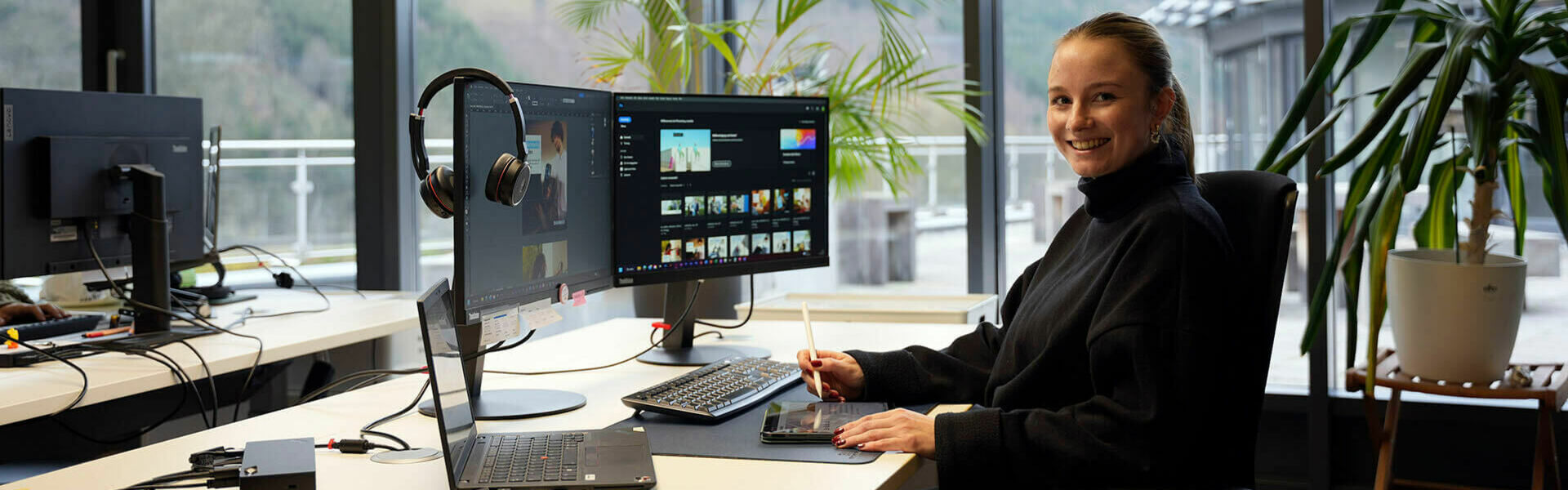 Eine junge Frau sitzt lächelnd an einem Schreibtisch in einem modernen Büro mit großen Fenstern. Vor ihr stehen zwei Monitore, ein Laptop und ein Grafiktablett. Im Hintergrund sind Pflanzen und eine Terrasse zu sehen.